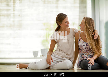 Frau legt den Arm um die Schultern ihrer halbwüchsigen Tochter und sie anlächeln, während er auf dem Boden, wie sie für ein Portrait darstellen. Stockfoto