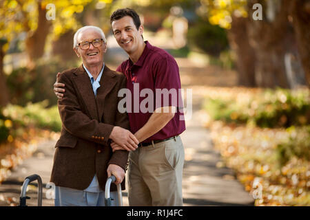 Portrait Of Happy älterer Menschen durch eine männliche Krankenschwester unterstützt, während sie seine Gehhilfe in einem Park. Stockfoto