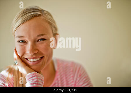 Fröhliche junge Frau lächelt, als sie ihr Kinn auf ihre Hand ruht. Stockfoto