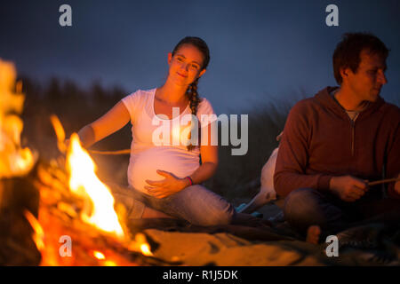 Schwangere junge Frau genießt toasten Marshmallows über einen Strand mit Lagerfeuer in der Nacht. Stockfoto