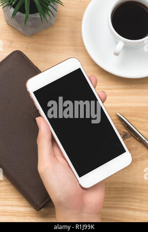 Männliche hand Smartphone mit Notebooks, Pen und Kaffeetasse neben auf Holz Tisch in morgen Zeit Stockfoto