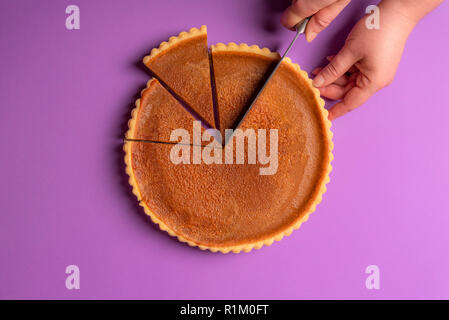Frau Hände Schneiden eine Kürbistorte auf einem violetten Hintergrund. Minimalistische kulinarische Konzept. Hausgemachten süßen Kuchen. Traditionelle Feiertage Dessert. Ansicht von oben. Stockfoto