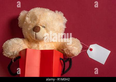 Teddybär Spielzeug in ein rotes Papier Beutel mit einem leeren weißen Tag, auf rotem Papier Hintergrund. Weihnachtsgeschenke einkaufen. Schwarzer Freitag. Stockfoto