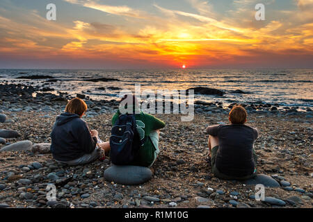 Dreiköpfige Familie (Mutter, Sohn, Tochter), die an einem steinigen Strand sitzt und den Sonnenuntergang in Westward Ho!, Devon, Großbritannien, beobachtet Stockfoto