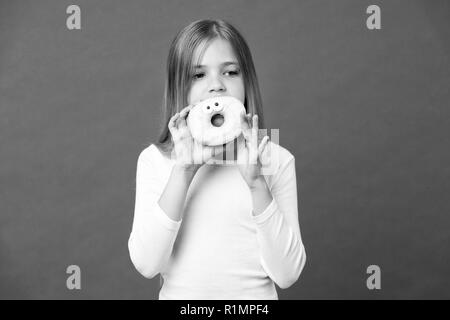 Mädchen, dass große Krapfen mit Puderzucker. Süßes, verglaste Bagel mit Augen. Kind essen lustig Dessert, süße Kindheit. Kind mit langen Haaren tragen weiße Outfit auf Violett Hintergrund isoliert. Stockfoto