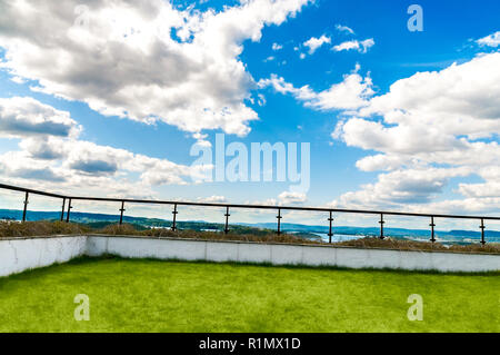 Eine hohe villa Terrasse mit modernen Glas Zaun und grünen Gras im Sonnenlicht mit einem top Luxus Panorama Blick auf das Meer und die Wolken am blauen Himmel Stockfoto