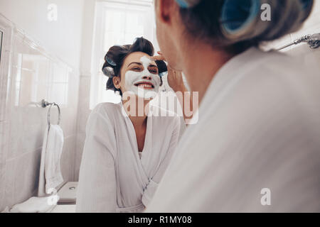 Lächelnde Frau im Bademantel ihr Gesicht zu Hause getan. Zwei Frauen im Badezimmer stehen mit Curly Rollen fixiert auf das Haar auftragen Gesicht Creme. Stockfoto