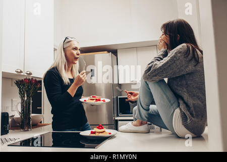 Ältere Frau cookies Essen aus einer Platte Standing in der Küche mit einer Frau. Zwei Frauen im Gespräch beim essen Snacks in der Küche. Stockfoto