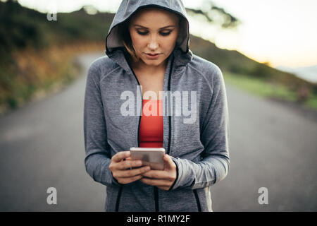 Fitness-Frau, die auf der Straße läuft und ein Kapuzenpullover trägt und auf ihr Handy blickt. Eine Frau, die ihr Handy benutzt und auf einer leeren Straße läuft. Stockfoto