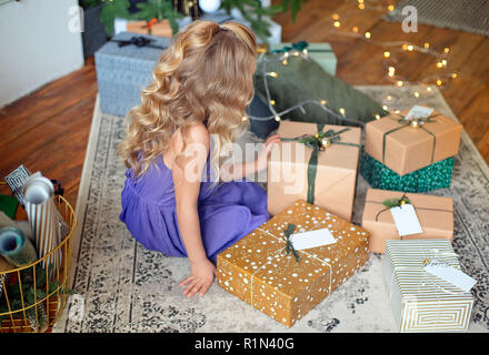 Ein kleines blondes Mädchen in bunten Kleid sitzt im Zimmer auf backround Geschenke. Urlaub Konzept. Mädchen auf dem Foto. Frohe Weihnachten, happy holiday Stockfoto