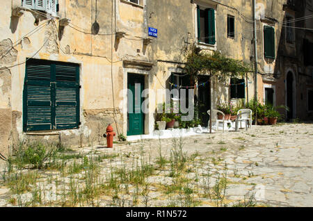 Blick auf einen Platz mit Gras überall wächst, in der Altstadt von Korfu in Griechenland, einige weiße Plastikstühle unter einer Pergola vor einem alten Stockfoto