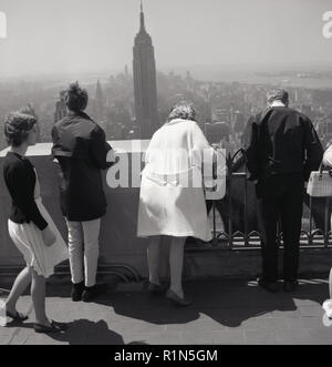 1950 s, historischen, "Nicht nach unten sehen!" ... Touristen stehen auf einem Balkon oder eine Aussichtsplattform auf der Spitze eines hohen Gebäude in Manhattan, New York, USA, Lean über den Rand einen Blick auf die Stadt unter einer der berühmtesten Wolkenkratzer der Stadt, das Empire State Building in der Ferne zu sehen ist. Stockfoto