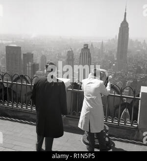 1950, historische, zwei männliche Touristen in regenmäntel stehen auf einem Balkon oder eine Aussichtsplattform auf der Spitze eines hohen Gebäude in Manhattan, New York, USA, eine mit einem Teleskop ein näherer Blick auf die Stadt und die berühmten Wolkenkratzer wie das Empire State Building in der Ferne zu sehen zu bekommen. Stockfoto