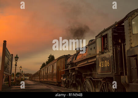 Niedriger Winkel, Seitenansicht Nahaufnahme des alten britischen Dampfzugs am Bahnsteig in leerem historischen Bahnhof am Ende des Tages mit glühendem Sonnenuntergang Abendhimmel. Stockfoto