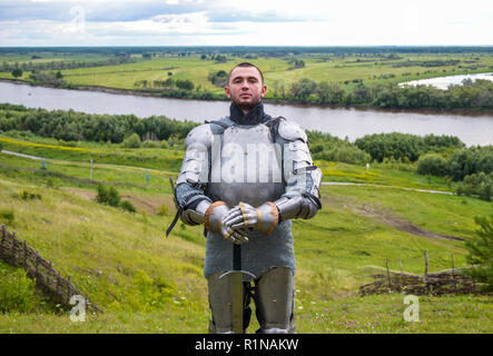 Ritter in Rüstung steht vor dem Hintergrund der native, offene Räume, Wälder und einen Fluss. Ritterlichen Rüstung und Waffe. Semi-antik Foto. Stockfoto