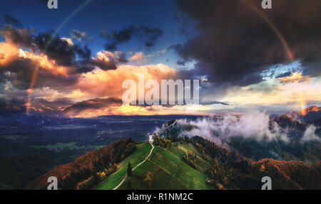 Jamnik, Slowenien - Luftaufnahme von Regenbogen über der Kirche St. Primoz in Slowenien in der Nähe von jamnik mit schönen Wolken und die Julischen Alpen im Hintergrund. Stockfoto