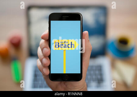 Ein Mann schaut auf seinem iPhone die zeigt die Am Strand logo, während saß an seinem Computer Schreibtisch (nur redaktionelle Nutzung). Stockfoto