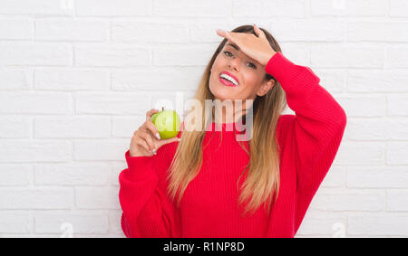 Junge erwachsene Frau über weiße Mauer essen frischen grünen Apfel mit Hand betonte am Kopf, mit Scham und Überraschung Gesicht schockiert, wütend und frustriert Stockfoto