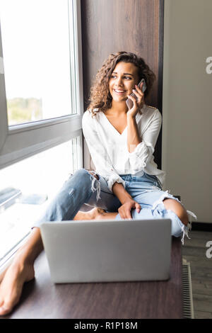 Fröhliche Frau sprechen Telefon und sitzen auf Fensterbank mit Laptop Computer zu Hause. Stockfoto