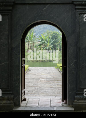 Ninh Binh, Vietnam. Abgebildet ist ein grauer Stein Tür führt zu einem Garten mit grünem Wasser und Bananenstauden. Stockfoto
