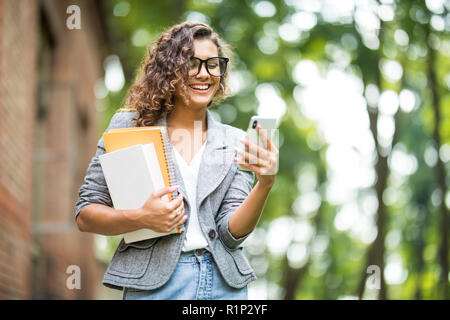 Einen fröhlichen Student mit mehrfarbigen Notebooks und eine Tasche ist zu Fuß rund um den Campus und am Telefon zu sprechen Stockfoto