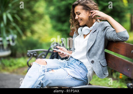 Nachdenkliche junge Schülerin im Kopfhörer. Entspannen und Musik hören, während Sie Kaffee trinken im Park. Lifestyle- und Technologiekonzept Stockfoto