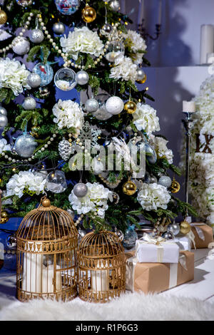 Leuchtenden Weihnachtsbaum mit Kugeln und Blumen, Geschenke und Kerzen in Käfigen unter es eingerichtet im Wohnzimmer. Weihnachten Abend. Urlaubsstimmung. Stockfoto