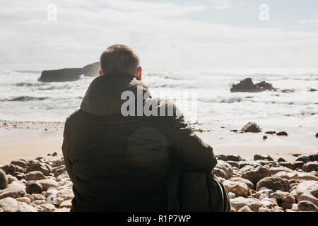 Ein Mann mit einem Rucksack oder ein Tourist oder ein Reisender in der Einsamkeit bewundert einen schönen Blick auf den Atlantik in Portugal. Suche nach Seele oder die Einheit mit der Natur. Stockfoto