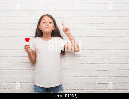 Junge hispanic Kid über weiße Mauer essen Rotes Herz lollipop Candy mit einer Idee oder Frage Zeigefinger mit glücklichen Gesicht überrascht, Nummer auf Stockfoto