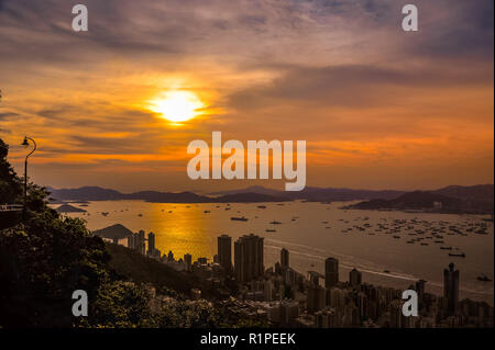 Sonnenuntergang über den Hafen von Hong Kong aus klitsch Lookout, Victoria Peak. Strahlend orange Sonne über entfernte Inseln mit Stadt Hochhäusern im Vordergrund. Stockfoto