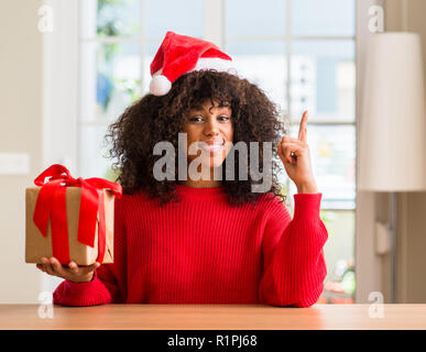 African American woman holding vorhanden tragen Christmas Red Hat mit einer Idee oder Frage Zeigefinger mit glücklichen Gesicht überrascht, Nummer eins Stockfoto