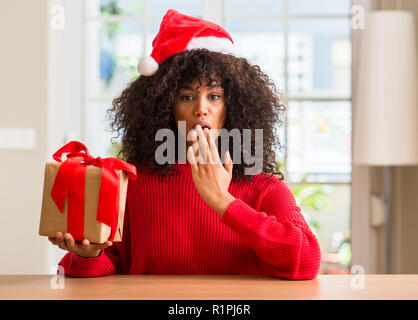 African American woman holding vorhanden tragen Christmas Red Hat decken den Mund mit der Hand mit Schande für den Fehler, den Ausdruck von Angst, in s Angst schockiert Stockfoto