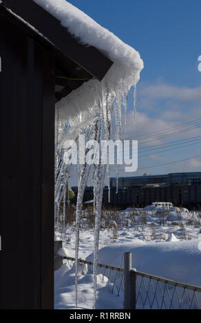 Lange transparente Eiszapfen auf dem Dach des Hauses mit Schnee und blauer Himmel im Hintergrund Stockfoto
