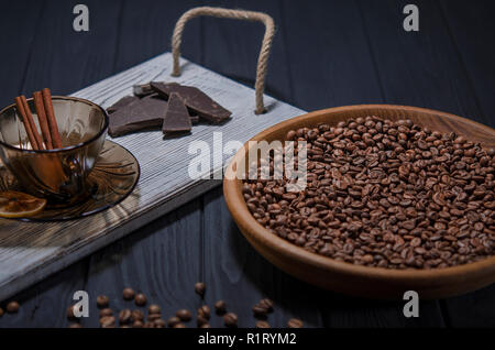 Geröstet duftende Körner schwarzen Kaffee liegen in einem braunen Holz- Platte, die auf einer schwarzen hölzernen Tisch steht und daneben steht ein weißes Holz- fach wit Stockfoto