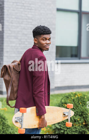 Lächelnden jungen Schüler mit Rucksack und Skateboard zu Fuß durch die Straße und die Kamera Stockfoto