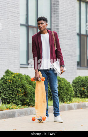 Junger student mit Skateboard und Notebooks stehen auf der Straße Stockfoto