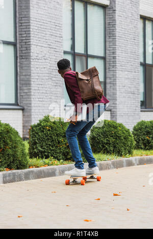Ansicht der Rückseite des jungen Studenten mit Rucksack und Notebooks reiten auf Skateboard auf der Straße Stockfoto