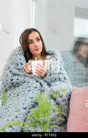 Schöne junge Frau auf der Fensterbank in Decke mit Tasse wärmenden Getränk zu Hause Schattendach Stockfoto