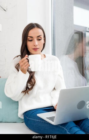 Schöne junge Freiberufler Kaffee trinken und Arbeiten mit Laptop auf dem Fensterbrett zu Hause Stockfoto