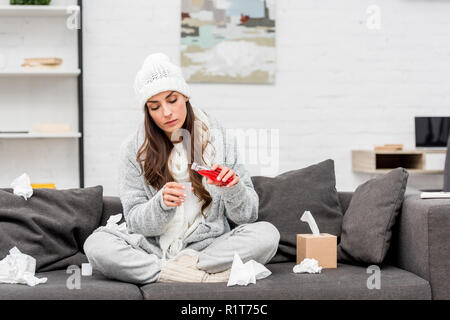 Erkrankte junge Frau in warme Kleidung gießen rot Hustensirup beim Sitzen auf der Couch zu Hause Stockfoto