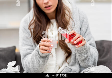 7/8 Schuß des Kranken jungen Frau in warme Kleidung gießen rot Hustensirup beim Sitzen auf der Couch zu Hause Stockfoto