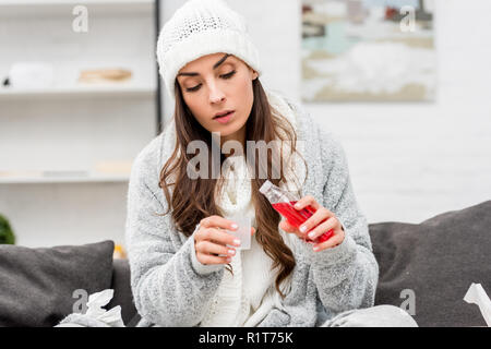 Kranke junge Frau in warme Kleidung gießen rot Hustensirup beim Sitzen auf der Couch zu Hause Stockfoto