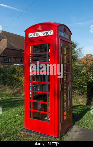 Altmodische rote Telefonzelle oder Kiosk auf der Straße in der Nähe des Village Hall im Dorf Rotherwick in Hampshire, Großbritannien, jetzt als eine Bibliothek verwendet Stockfoto