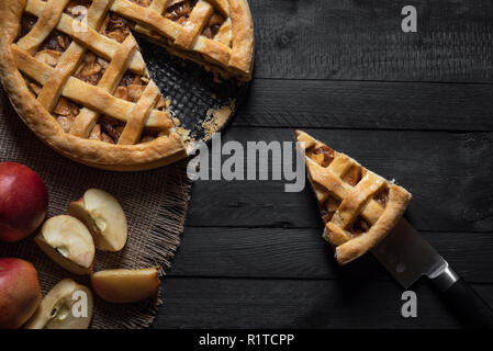 Blick von oben auf ein Stück Kuchen auf ein Messer auf einem urigen schwarz Tisch und die ganze Torte und Äpfel. Hausgemachte Desserts. Urlaub pie Kontext. Stockfoto