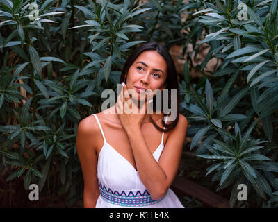 Schöne brünette Mädchen in weißem Kleid sitzt in der Nähe von grünen Bäumen Stockfoto