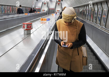 U-Bahn Pendler Zeit, junge Frau trägt ein Kopftuch und mit ihrem Smartphone als reist sie die Rolltreppe nach oben in der Londoner U-Bahn Stockfoto