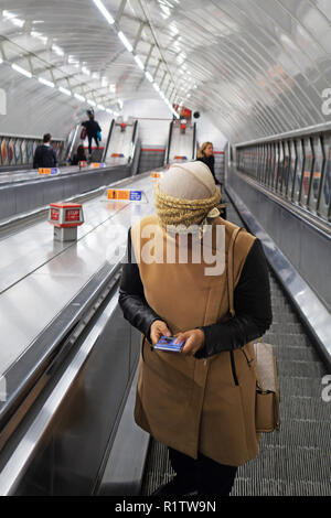 U-Bahn Pendler Zeit, junge Frau trägt ein Kopftuch und mit ihrem Smartphone als reist sie die Rolltreppe nach oben in der Londoner U-Bahn Stockfoto