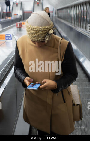 U-Bahn Pendler Zeit, junge Frau trägt ein Kopftuch und mit ihrem Smartphone als reist sie die Rolltreppe nach oben in der Londoner U-Bahn Stockfoto