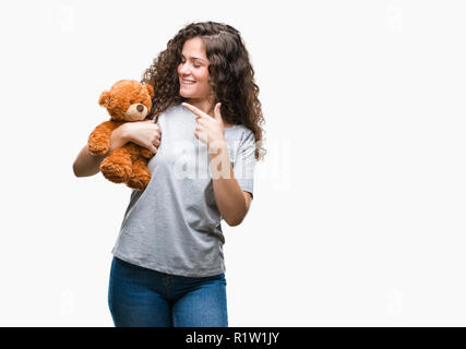 Junge brünette Mädchen, dass Teddybären über isolierte Hintergrund sehr glücklich zeigte mit Hand und Finger Stockfoto