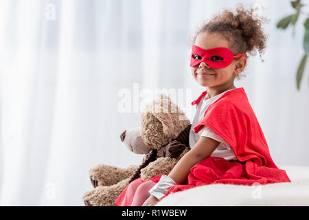 Adorable kleine afrikanische amerikanische Kind in Superhelden Kostüm und Maske mit Teddybär lächelnd an Kamera Stockfoto